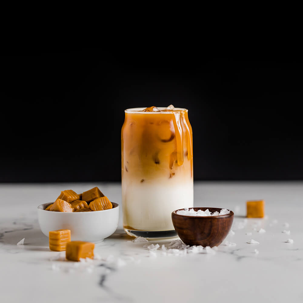 An iced caramel drink, a bowl of caramels, and a bowl of salt are on a white surface.