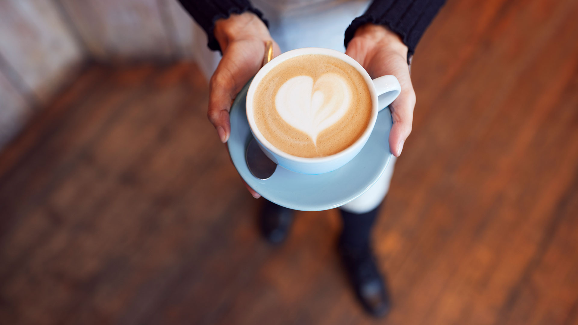 Hands hold a light blue cup of latte with a heart design in the foam, over a wooden floor.