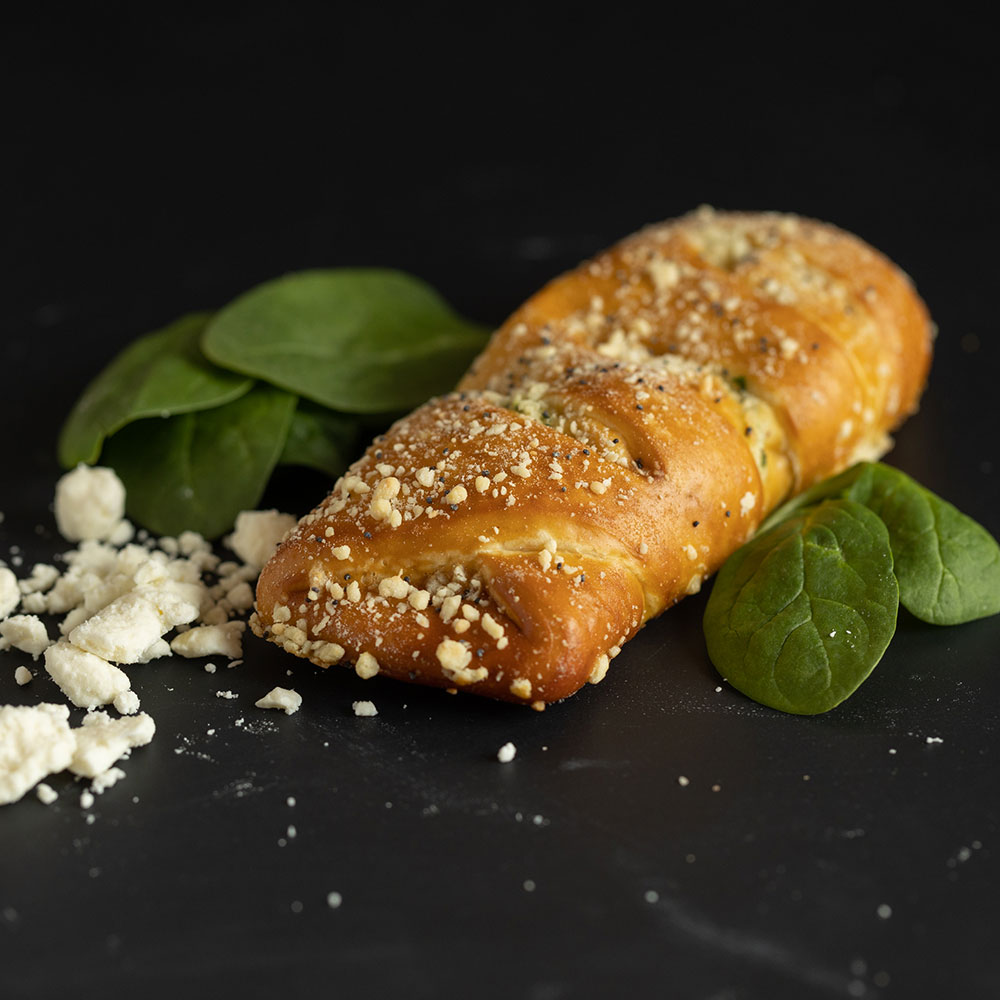 A golden-brown pretzel stick topped with crumbled feta cheese and spices, next to fresh spinach leaves.