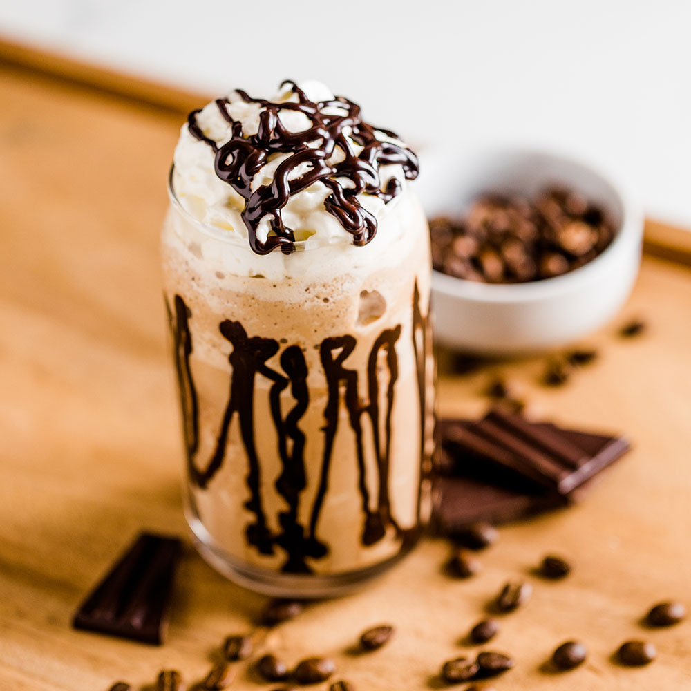 A glass of mocha with whipped cream and chocolate drizzle, surrounded by chocolate pieces and coffee beans.