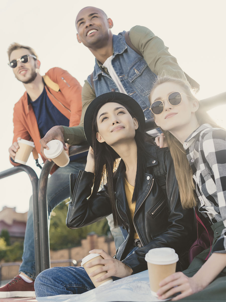 Four friends with diverse appearances are smiling and holding coffee cups on a sunny day.