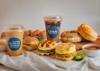 An iced coffee with "Canal Coffee Co." and "Flavor Favors the Traveled" text, breakfast sandwiches, bagels, and eggs.