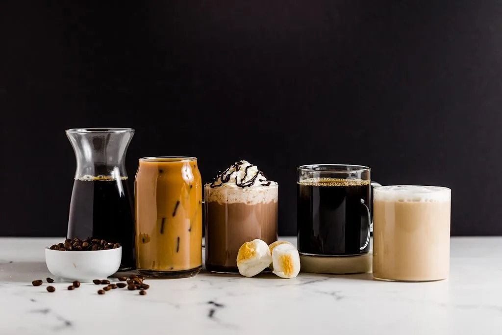 A row of coffee drinks: a cold brew carafe, iced latte, mocha, black coffee, and a latte on a marble sruface.