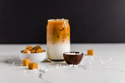 A clear glass with an iced latte, featuring a layer of milk and coffee, next to bowls of caramel candies and sea salt.