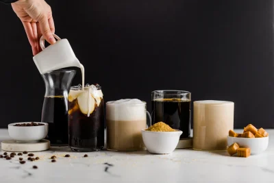 A hand pours white cream into an iced coffee next to coffee beans, sugar, and caramel candies on a white table.