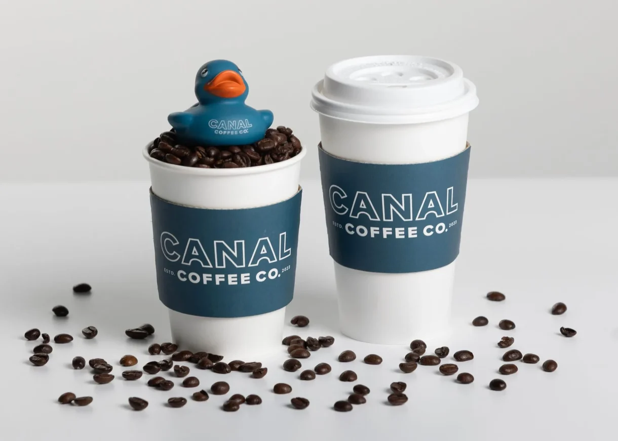 Two Canal Coffee Co. cups, one filled with coffee beans and a rubber duck, surrounded by spilled beans.