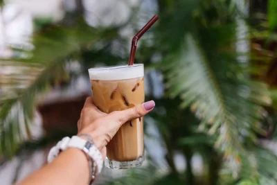 A hand with pink fingernails and a white watch holds an iced coffee with a brown straw against blurred leaves.