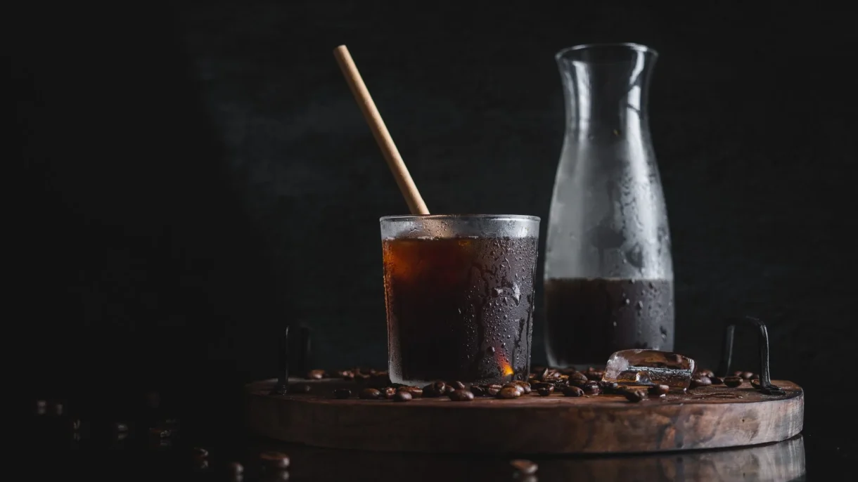 Cold brew coffee in a sweating glass with a straw, a carafe, and coffee beans on a dark wooden tray.