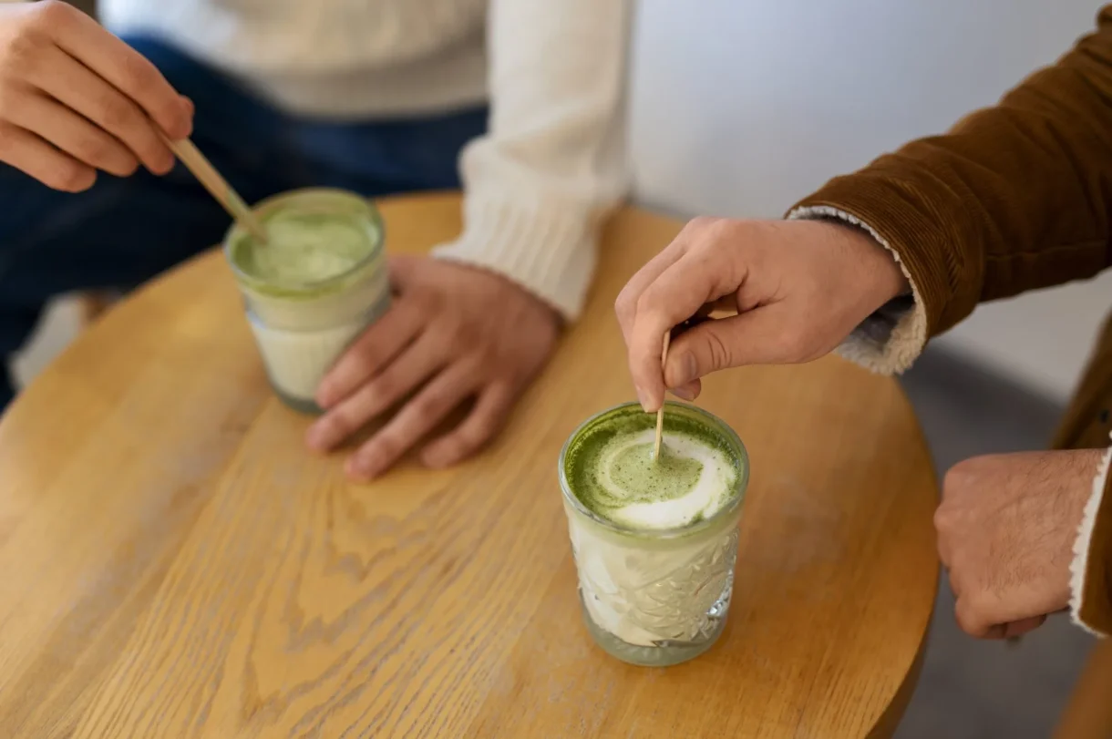 Two people stirring green matcha drinks with wooden spoons at a wooden table, hands visible wearing brown and white sleeves.
