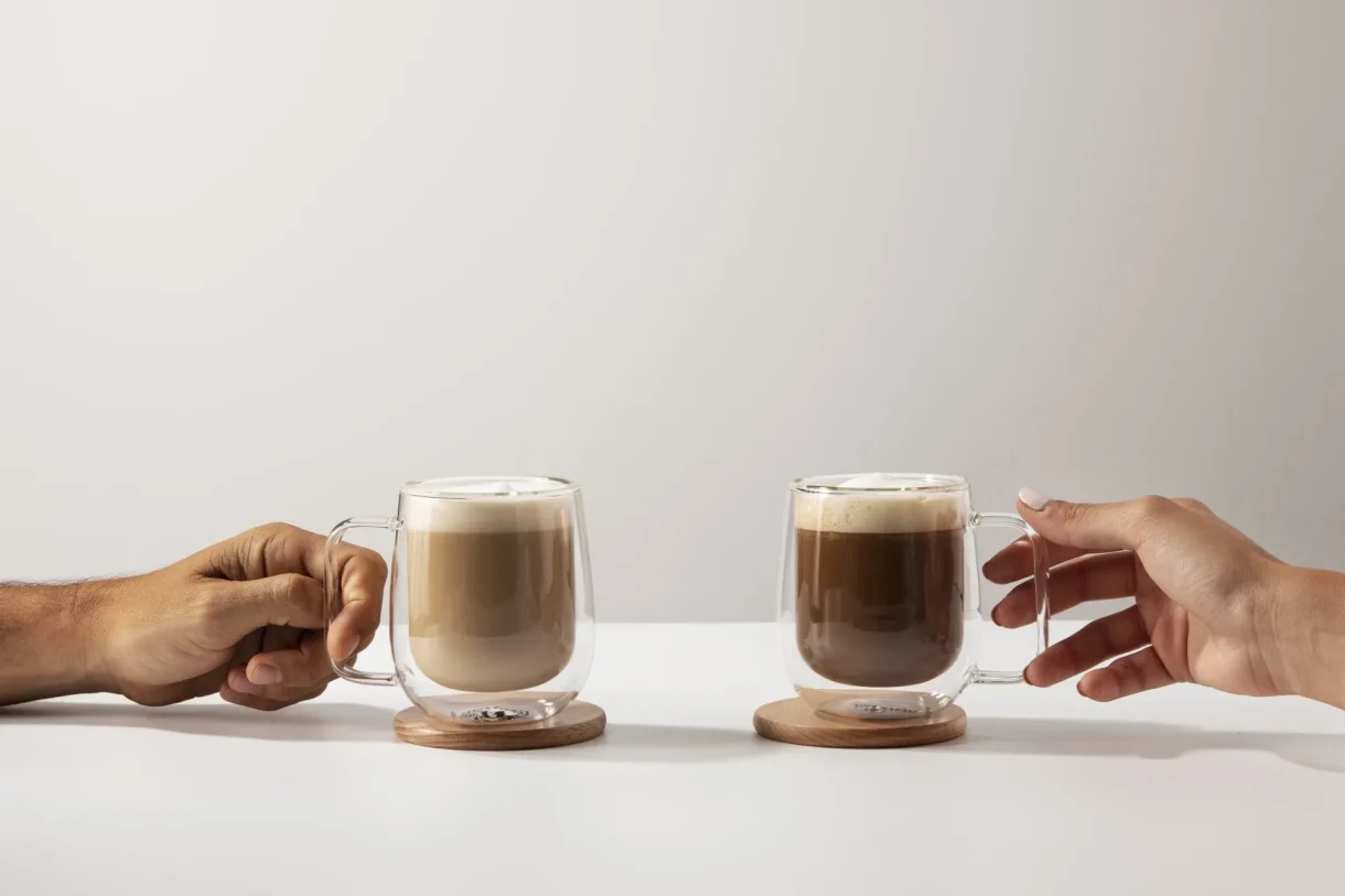 Two hands hold double-walled glass mugs of foamed coffee, one light latte and one dark cortado, on wood coasters.