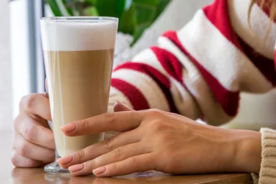 Woman in red and white striped sweater holds a layered latte in a tall glass at a table with a pastry nearby.