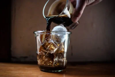 Dark coffee pours from a small glass into a tumbler filled with ice cubes and a large spherical ice ball on a wood table.