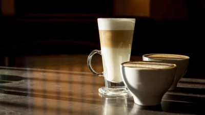 A tall clear glass of layered latte macchiato next to two white ceramic cups of cappuccino on a sunlit wooden table.