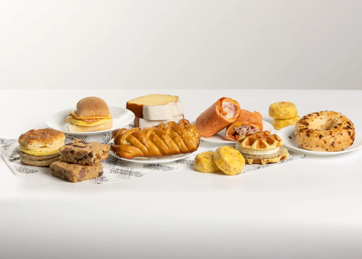 Assorted Canal Coffee Co food items including sandwiches, wraps, a bagel, pastries, and egg bites on a white surface.