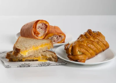 A sliced turkey and cheese wrap, a grilled cheese sandwich, and a braided spinach pastry on white plates and branded paper.