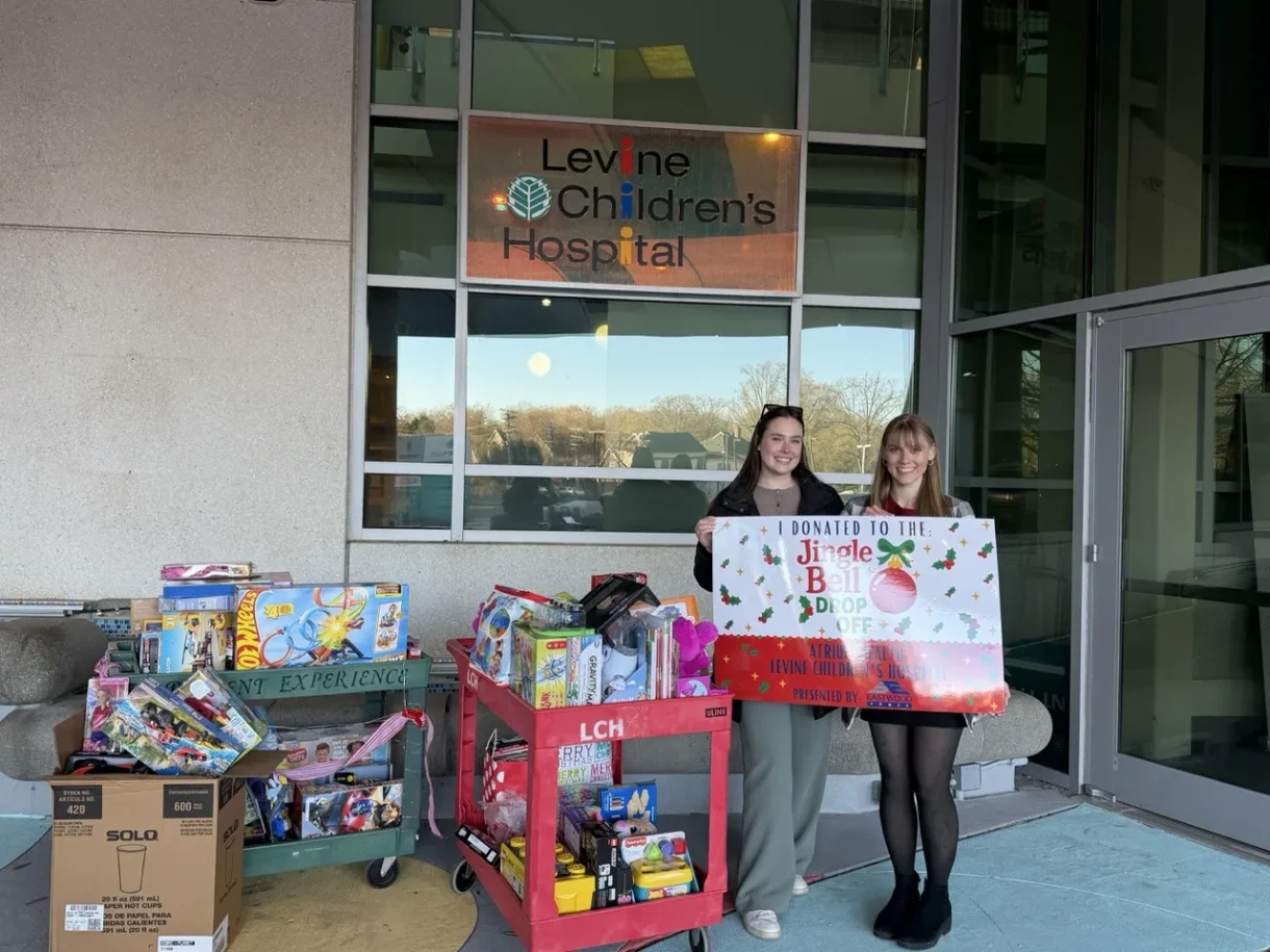 Two women with donated toys and a Jingle Bell Drop Off sign outside Levine Children’s Hospital.