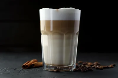 Layered latte in a glass with foam, coffee beans, and cinnamon sticks.