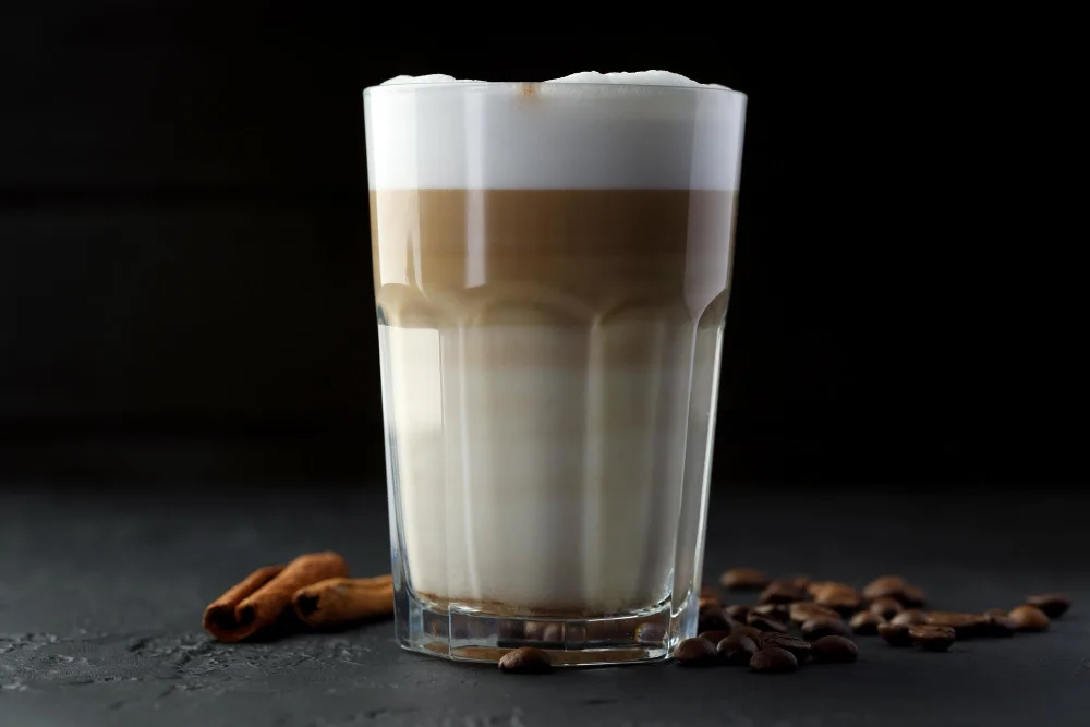 Layered latte in a glass with foam, coffee beans, and cinnamon sticks.