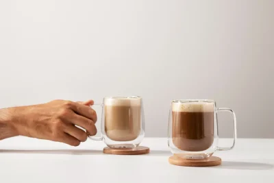 Hand holding a glass mug of coffee beside another coffee drink.