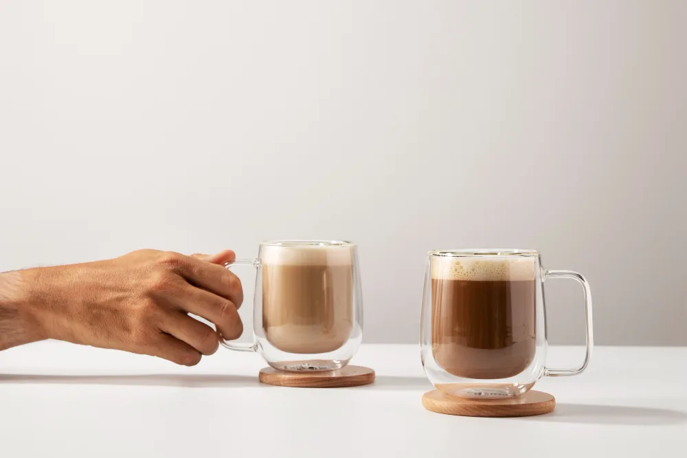 Hand holding a glass mug of coffee beside another coffee drink.