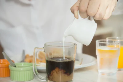 Milk being poured into black coffee in a clear glass mug.