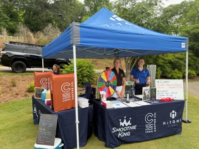 Canal Coffee Co. and HiTONE Fitness booth at an outdoor event. Two woman stand behind a desk with paper and a prize wheel.