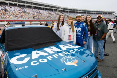 Group of six people posing beside a Canna Coffee Co. race car at a racetrack.