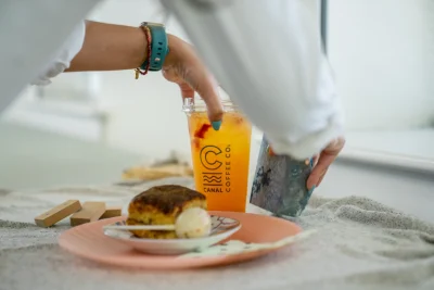 Person picking up an iced Canal Coffee drink next to a pastry.